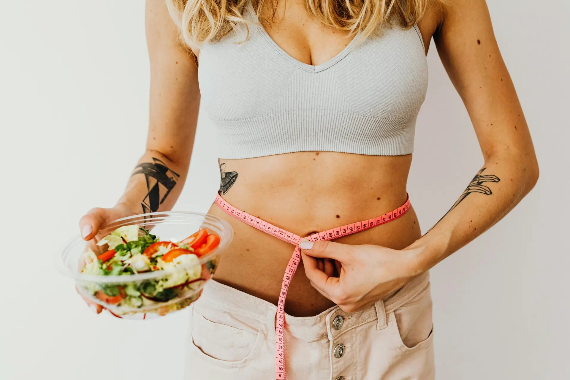 Woman measuring waistline while holding a fresh vegetable salad, emphasizing healthy living.